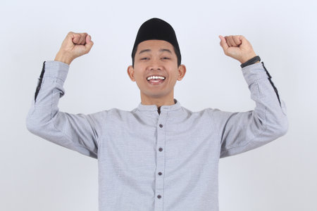 Joyful young Asian Muslim man in traditional clothes celebrating victory. Happy expression for Ramadan, Eid al-Fitr, or success concept. Isolated portrait.の写真素材