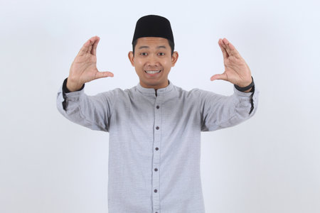 Cheerful Indonesian man in traditional peci and koko shirt making a holding gesture. Perfect for Ramadan or Eid Mubarak greetings and promotions.の写真素材
