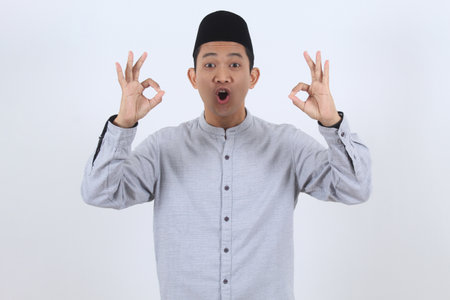 Excited young Asian Muslim man in traditional peci cap shows a double OK sign. Concept of approval, satisfaction, success, or Ramadan celebration.の写真素材