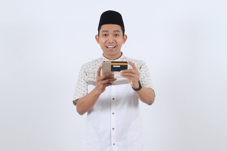 Happy young Asian Muslim man holds a smartphone and credit card for online payment or e-commerce...