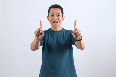 Portrait of a cheerful young Asian man smiling happily, pointing up with both index fingers. Concept for ideas, solutions, and positive announcements.の写真素材