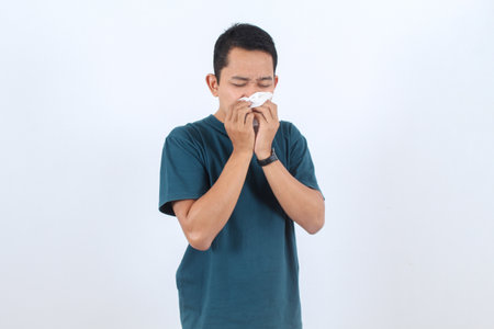 Unwell young man suffering from a runny nose, sneezing into a napkin. Concept of respiratory illness, rhinitis, or hay fever. Isolated portrait.の写真素材