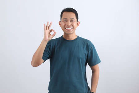 Portrait of a friendly young Asian man in a casual t-shirt making an OK sign. Symbol of approval, satisfaction, success, and positive agreement.の写真素材