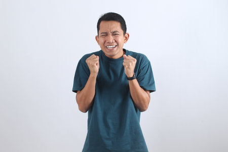 Portrait of an expressive young Asian man clenching his fists, feeling overjoyed and successful or...