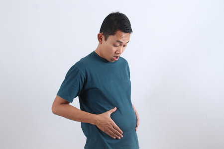 Unhappy man holds his bloated belly in shock. Concept for overeating, indigestion, weight gain, poor...