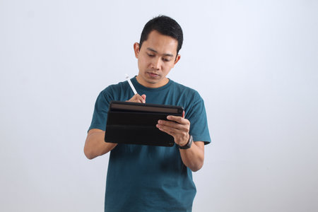 Focused Asian man in casual wear using a stylus on a tablet. Digital artist, designer, or student working, drawing, or signing a contract online.の写真素材