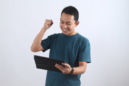 Overjoyed Asian man celebrates a business victory or online win, looking at his tablet with a happy smile and a fist pump. Isolated on white.の写真素材