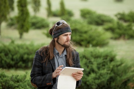 Young hipster fashion guy with the tablet sitting in the park in summer weather and plays.の写真素材
