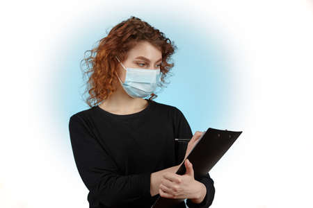 Young girl with a protective mask on her face with a notebook and a pen in her hands writes down the...