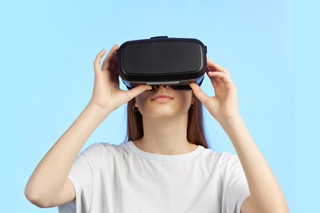 Young teen girl in a virtual reality helmet isolated on a blue background plays a 3d computer game.