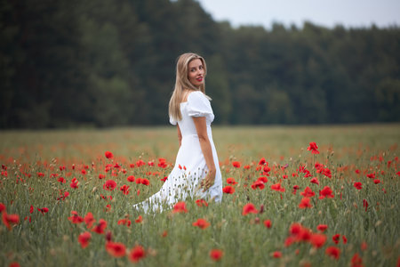 A young slim woman in a white costume walks in a flower field with poppies in summer.の写真素材