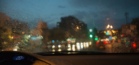 A photograph of the lights of traffic lights and car headlights through the windshield of the car during heavy rain. Blurred colored traffic lights and street lights during rain. Drops and splashes on the windshield of the car.の写真素材