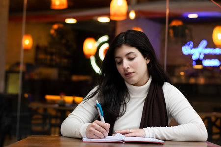 Seated woman writing. restaurant in the background. Thoughtful woman writing in a notebook. Woman sitting in a restaurantの写真素材