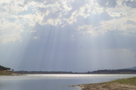 Scenic view of sun rays filtering through thick clouds, casting light over a calm lake and distant shoreline. Peaceful and serene atmosphere, perfect for illustrating nature, weather, tranquility, meditation, and environmental concepts.の写真素材