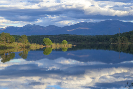 A dramatic, tranquil scene of a lake or reservoir perfectly reflecting dark mountains and cloudy, blue skies. The clean reflection and moody atmosphere are ideal for nature, peace, and travel themes.の写真素材