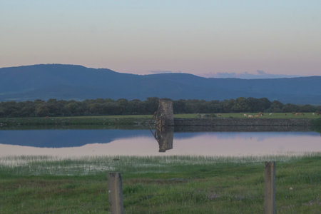 Peaceful dusk landscape featuring a calm lake reflecting an old stone ruin. Cows graze in the green meadow behind with blue layered mountains under a soft pink and purple gradient sky.の写真素材