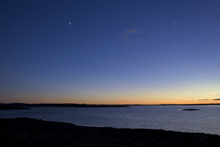 Wide view of a calm reservoir at twilight. The sky shows a smooth gradient from orange to deep blue with a small crescent moon. Dark silhouettes of the shore frame the water in the foreground.の写真素材