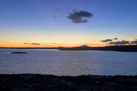 Wide horizontal landscape of the reservoir at sunset. The calm water perfectly reflects the dramatic pink, orange, and blue gradient of the sky over the distant silhouetted hillsの写真素材