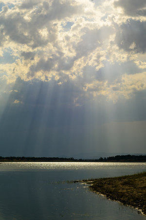 Wide majestic landscape of the Gabriel y Galán reservoir. Dramatic dark clouds filter sunlight creating visible crepuscular rays that pierce the sky and illuminate the water and distant hillsの写真素材