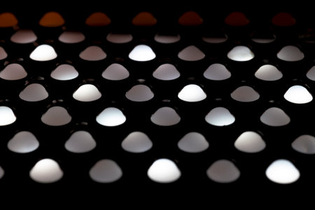 Abstract close up of light shining through the holes of a metal kitchen grater. The backlit pattern creates glowing white and orange semi circles against a dark silhouetted background.の写真素材