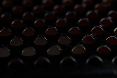 Abstract macro shot of a perforated metal surface (grater) taken from a different angle in low light. The holes create a dark, minimal pattern of glowing light and intense shadow, suitable as a digital background.の写真素材