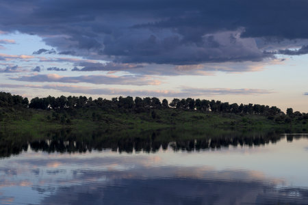 Calm lake reflecting trees and dramatic sky with soft twilight colors creating a serene natural landscapeの写真素材