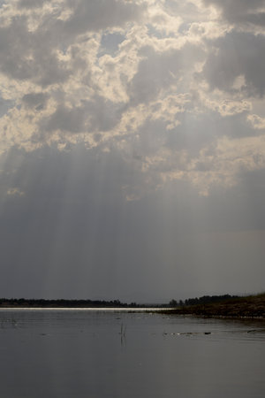 Dramatic sun rays breaking through dense clouds over a calm lake creating a peaceful atmospheric natural landscapeの写真素材