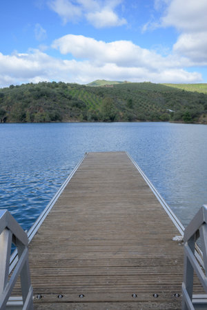 Wooden dock at La Pesga in Caceres overlooking calm reservoir waters with green hills and blue sky...
