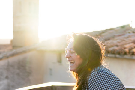 Brown-haired woman with glasses laughing out loud at sunset in rural environment with a bell tower in the backlit backgroundの写真素材