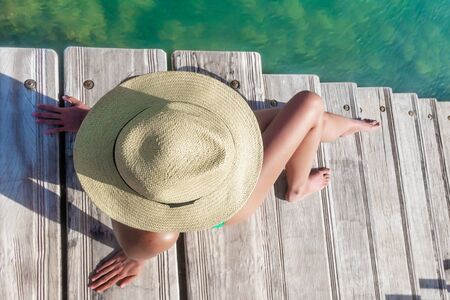 Svelte Caucasian woman with hat seen from above sitting relaxed on wooden stairs by the sea.の写真素材