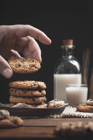 Hand holding chocolate chip cookie with a glass and a bottle of milk on a wooden base, dark background. Sweet food concept.の写真素材