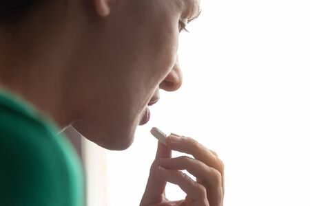 Young Caucasian woman in green sweater taking a pill in front of the window. Medicine and health...