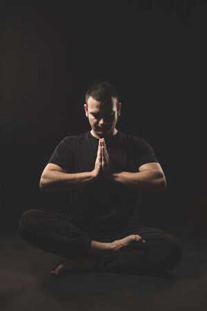 Young Caucasian man sitting with hands in namaste gesture with black clothes and black background.の写真素材