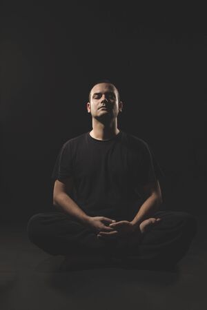 Young Caucasian man sitting in meditation with black clothes and black background.の写真素材