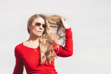 Caucasian blonde woman with red sweater and sunglasses posing outdoors.の写真素材