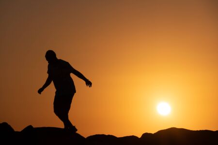 Man silhouette walking on rocks, orange sky at sunset.の写真素材