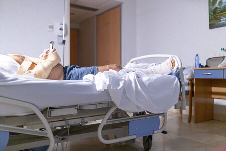 Patient lying in hospital bed with broken leg looking at the cell phone. Hospitalization and medical care concept.の写真素材
