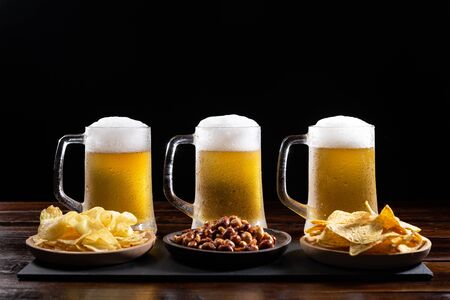 Three ice-cold beer mugs and plates with appetizers on wooden base on black background.の写真素材