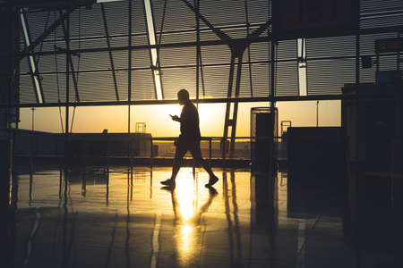 silhouette of an unrecognizable man staring at his mobile phone as he walks through the airport terminal at sunrise.の写真素材
