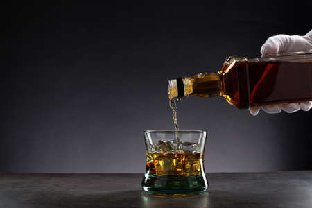 White gloved hand pouring whiskey into a glass with ice on dark background, with copy space.