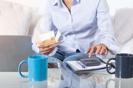 Close up women's hands doing accounting with calculator and bills in hand, on the couch at home.