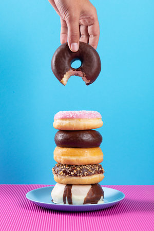 Hand holding bitten donut and stack of different donuts on pink and blue background.の写真素材