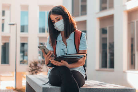 Young brown-haired girl with a mask checking on her mobile phone the notes of the university to studyの写真素材
