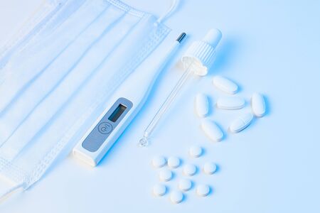 electronic thermometer, protective mask, pipette and tablets pills lie on a white background with blue illuminationの写真素材