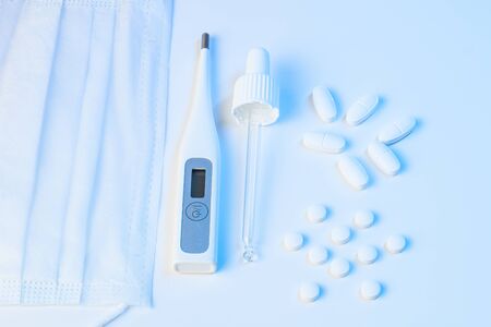 electronic thermometer, protective mask, pipette and tablets pills lie on a white background with blue illuminationの写真素材