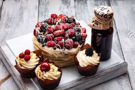pancake cake with a cap of berries drenched in white chocolate on a wooden white table with a few Belgian muffins and a bottle of syrupの写真素材
