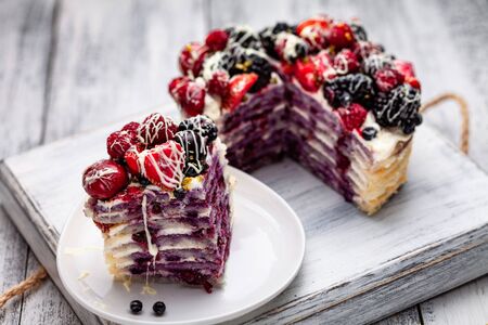 a piece of pancake cake with a cap of berries doused with white chocolate on a plate with Belgian muffins and a bottle of syrup on a tray on a white wooden tableの写真素材