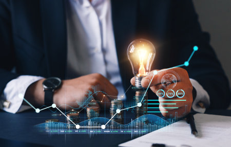 Businessman hand holding light bulb and coins stack with graph and chart. Business idea and innovation concept.の写真素材