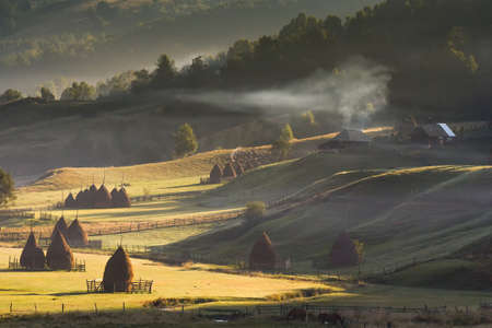 Beautiful rural mountain landscape in the morning light with fog, old houses and haystacks, Fundatura Ponorului, Hunedoara County, Romaniaの写真素材