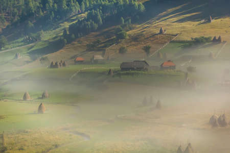Beautiful rural mountain landscape in the morning light with fog, old houses and haystacks,...
